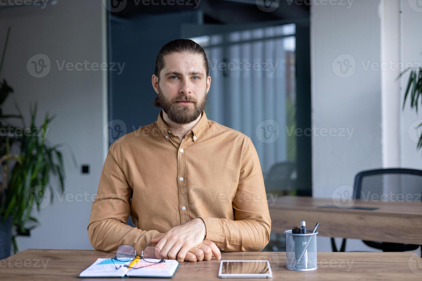 A man with a beard sits at his desk with a tablet and notebook, in a modern office setting. He has a serious look and is staring ahead. photo