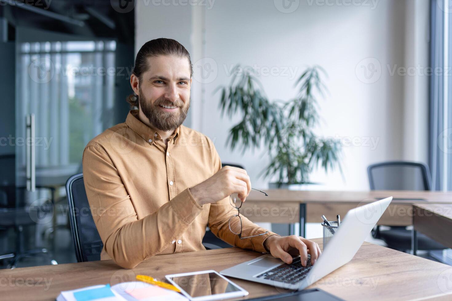 A smiling businessman at his desk, holding glasses, works on his laptop in a modern office setting. The man looks directly at the camera, radiating confidence. photo