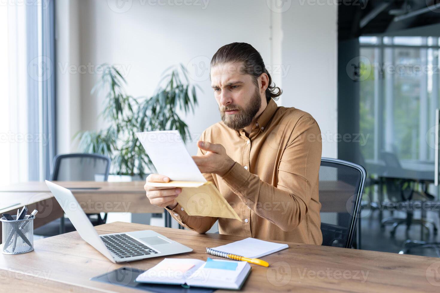 A focused businessman is looking at documents in his hands, possibly analyzing data at his desk during work hours in his office. photo