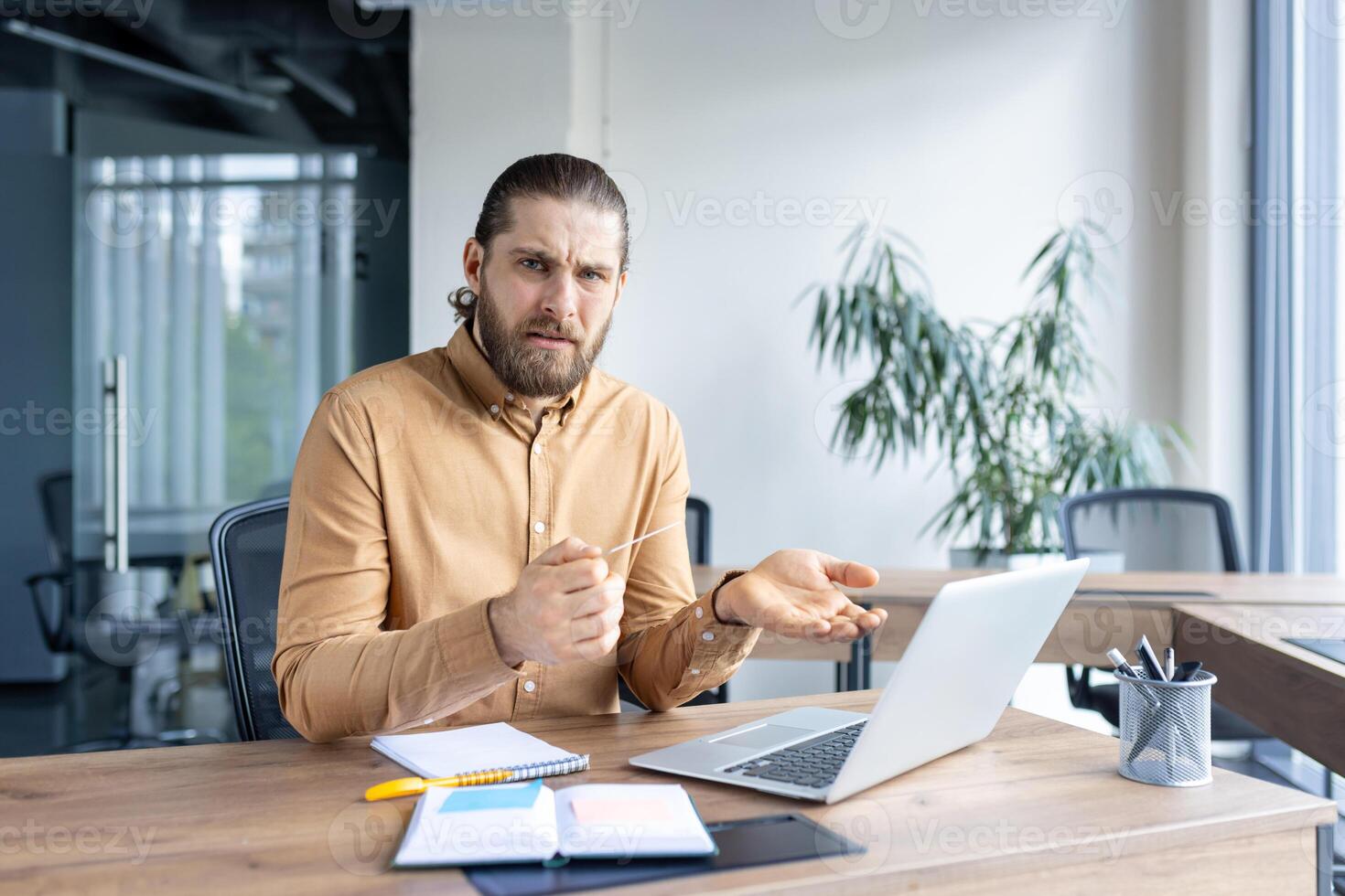 A bearded man is looking at his laptop and expressing disbelief or confusion during a conference in a modern office setting. photo