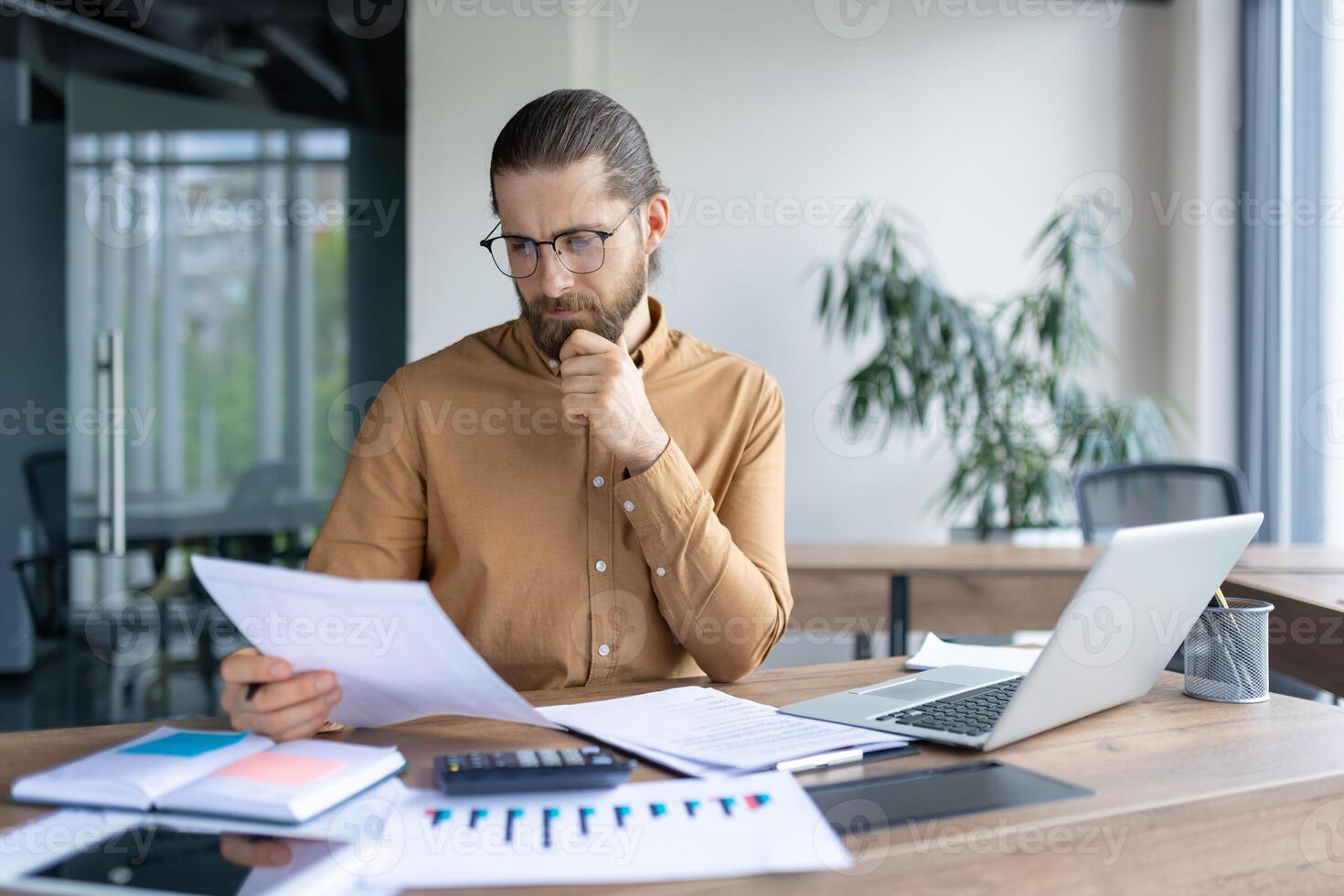 Bearded businessman in glasses examines documents at his desk with a laptop, calculator, and charts, thoughtfully analyzing information. photo