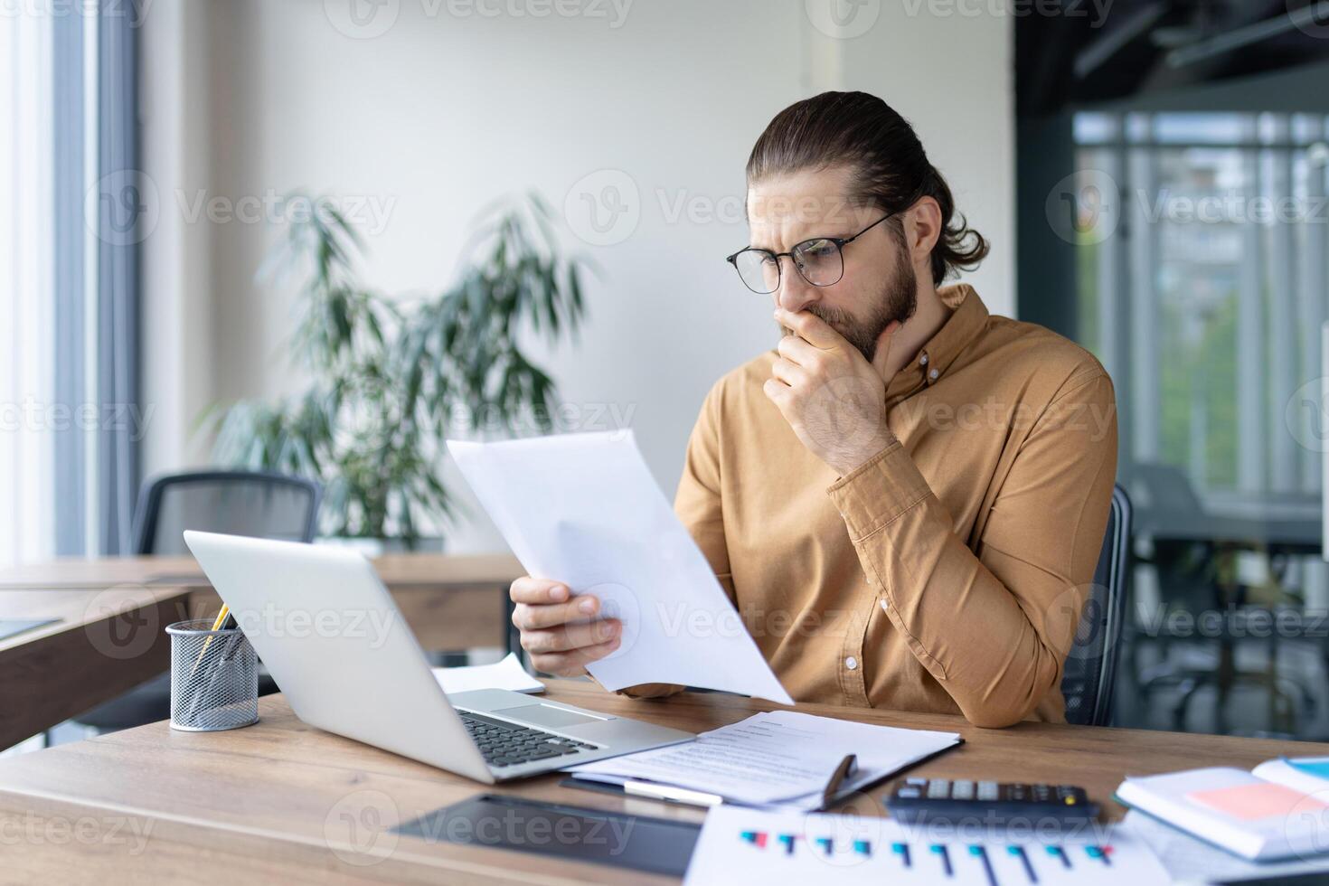 A focused professional reviews documents at his desk in an office setting, pondering the information. Laptop, papers, and calculator are visible. photo