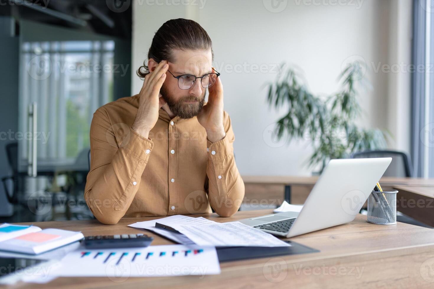 A stressed businessman massaging his temples while looking at a laptop in his office, surrounded by charts and financial documents. photo