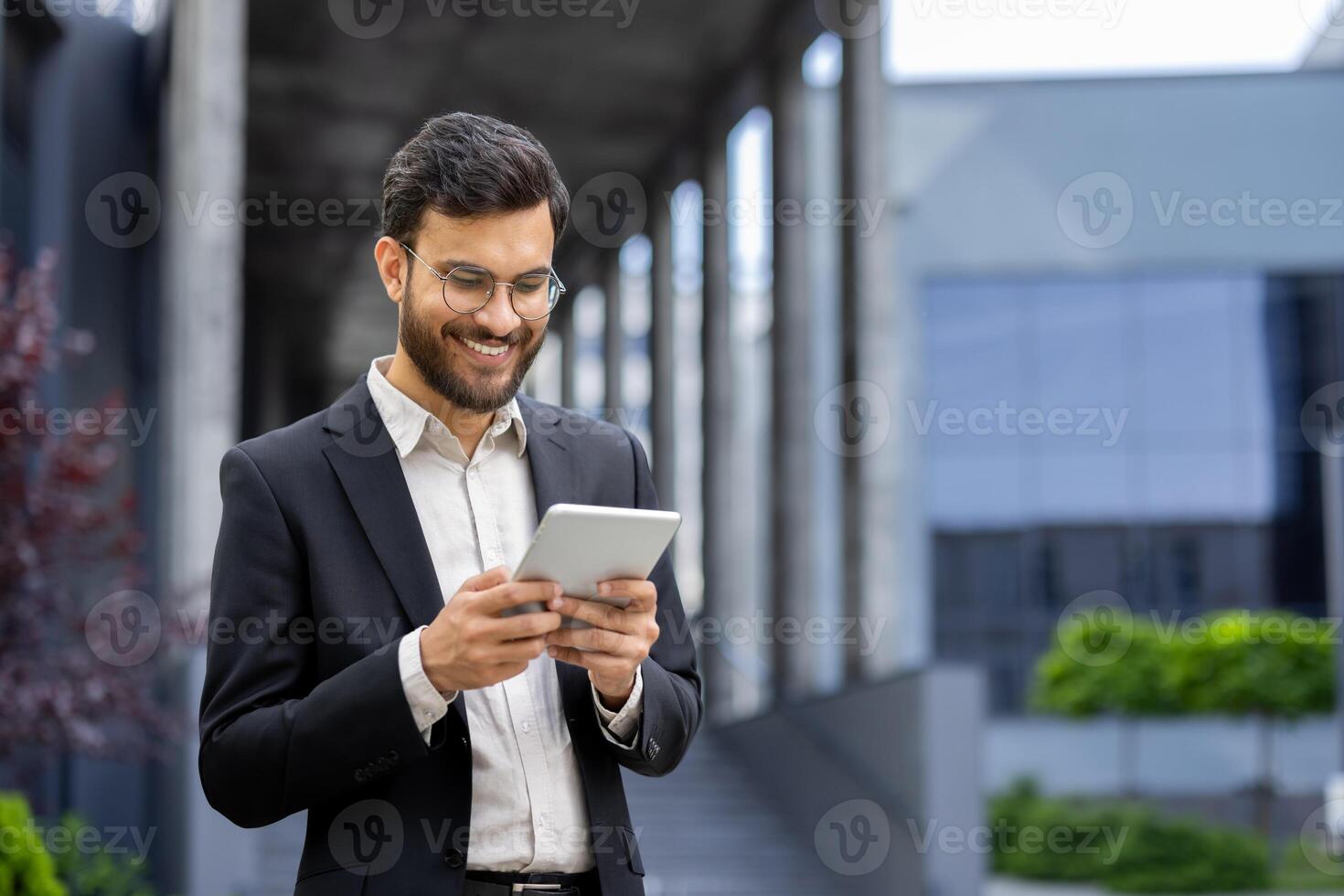 A smiling businessman in a suit works on his tablet while standing outside a modern office building on a sunny day. photo