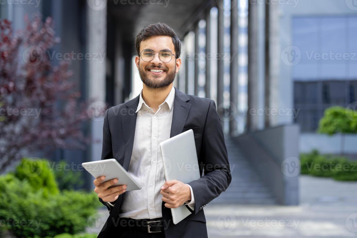 A smiling businessman holds a tablet and a laptop, showing his modern, digital tools. The man is well-dressed with glasses in front of a modern building. photo