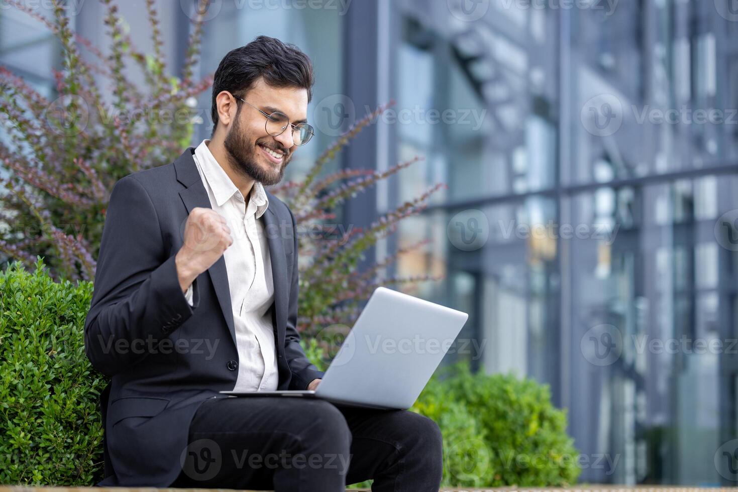 A businessman in a suit celebrates a successful project result while working on his laptop outdoors near an office building. photo