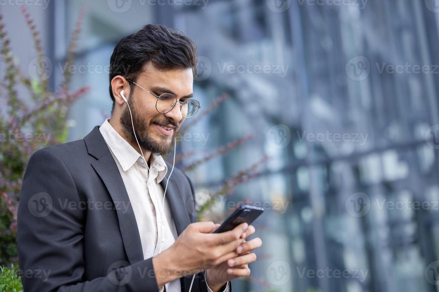 A young businessman in a suit smiles as he looks at his phone and listens with earphones near a modern building. photo