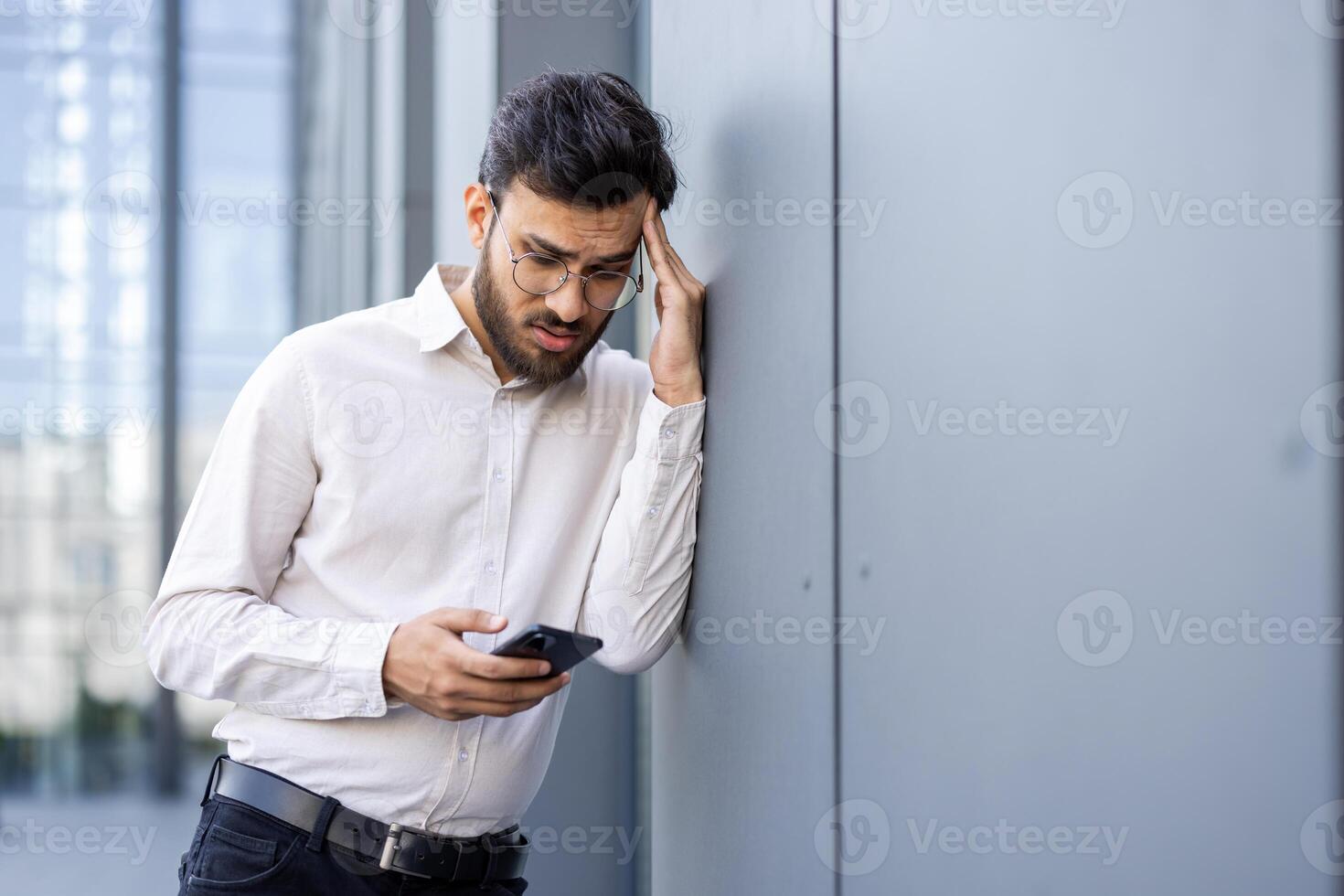 A concerned businessman in glasses looks at his phone while leaning against a building wall outside. He appears stressed. photo