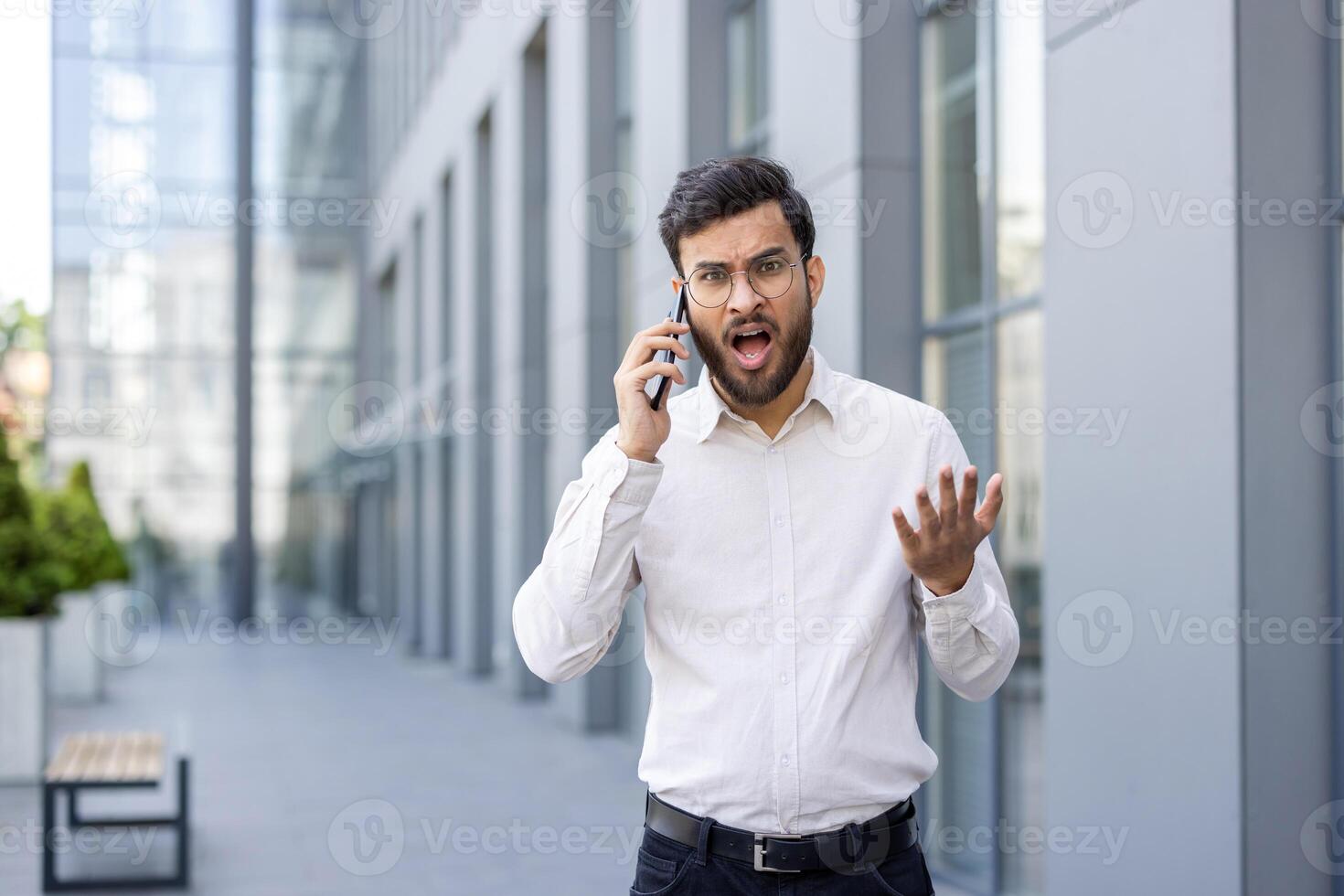 Anxious man is on the phone, expressing frustration with an open mouth and raised hand, outdoors. photo