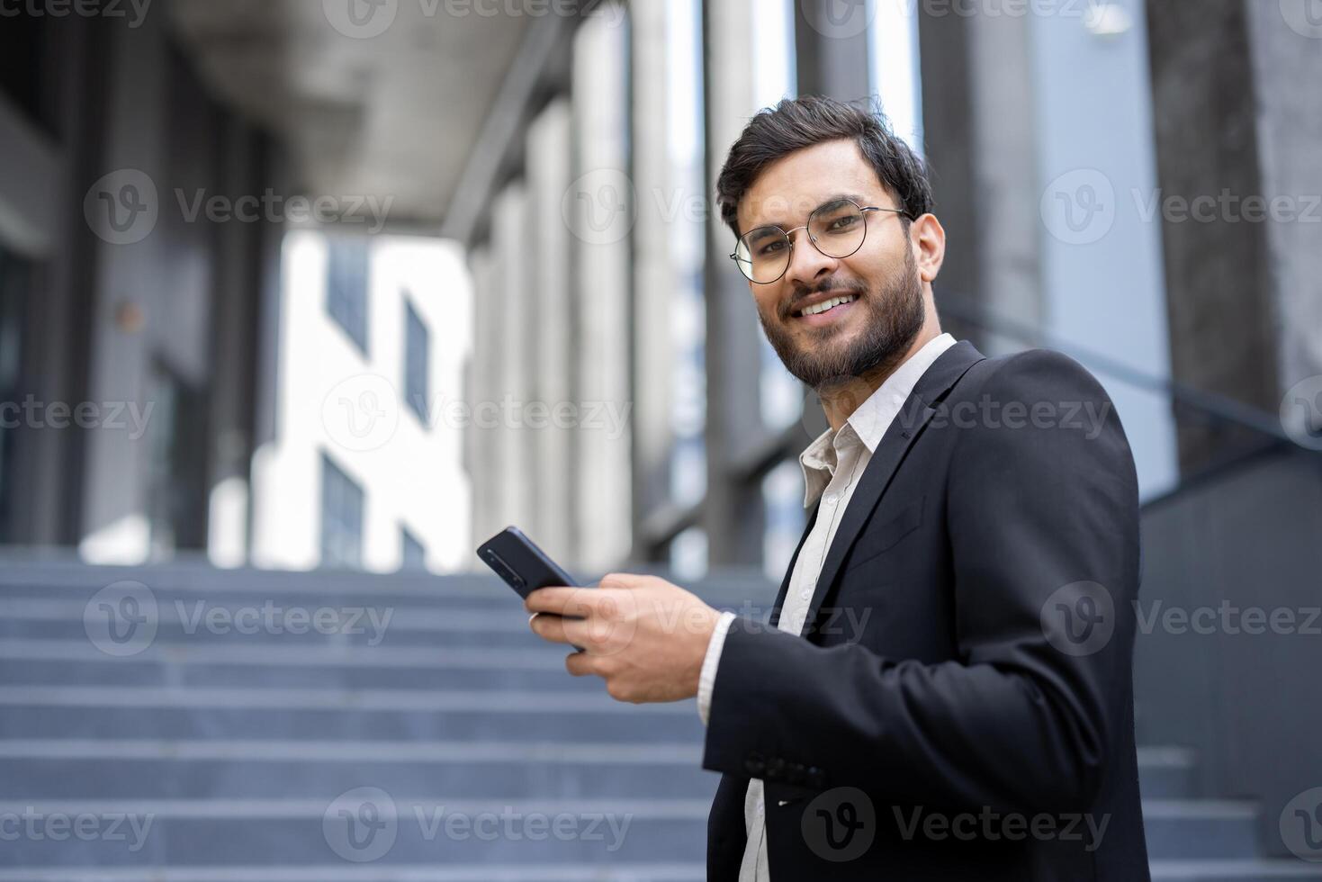 A smiling businessman, dressed in a suit, holding a phone, looks at the camera while standing outside a modern building. photo