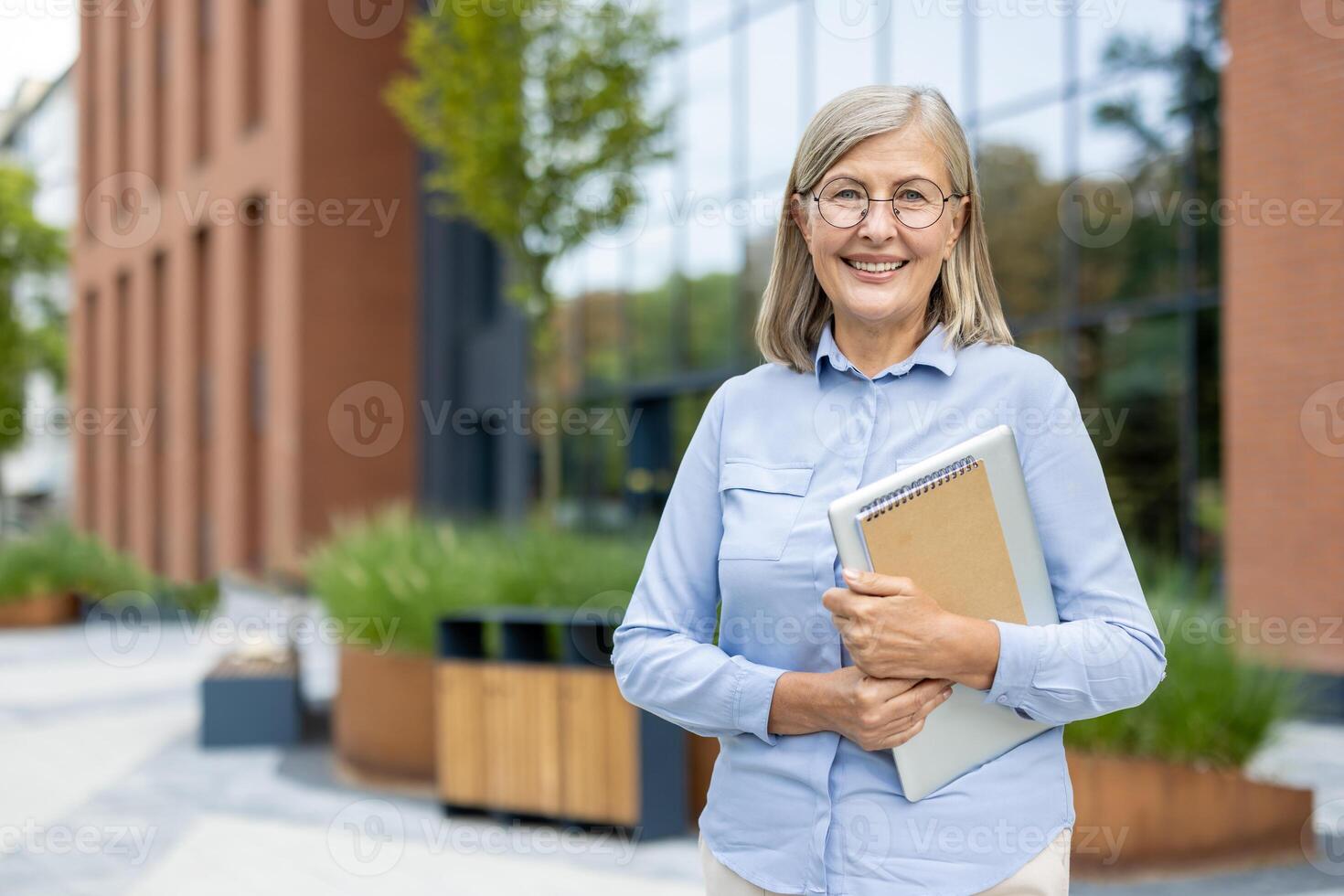 A smiling, mature professional woman stands confidently outside an office building holding a laptop and notebook, showcasing her preparedness. photo