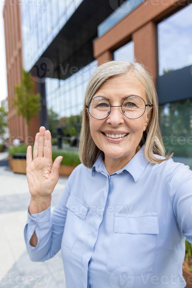 A friendly senior woman waves in front of a modern office building, smiling and welcoming. photo