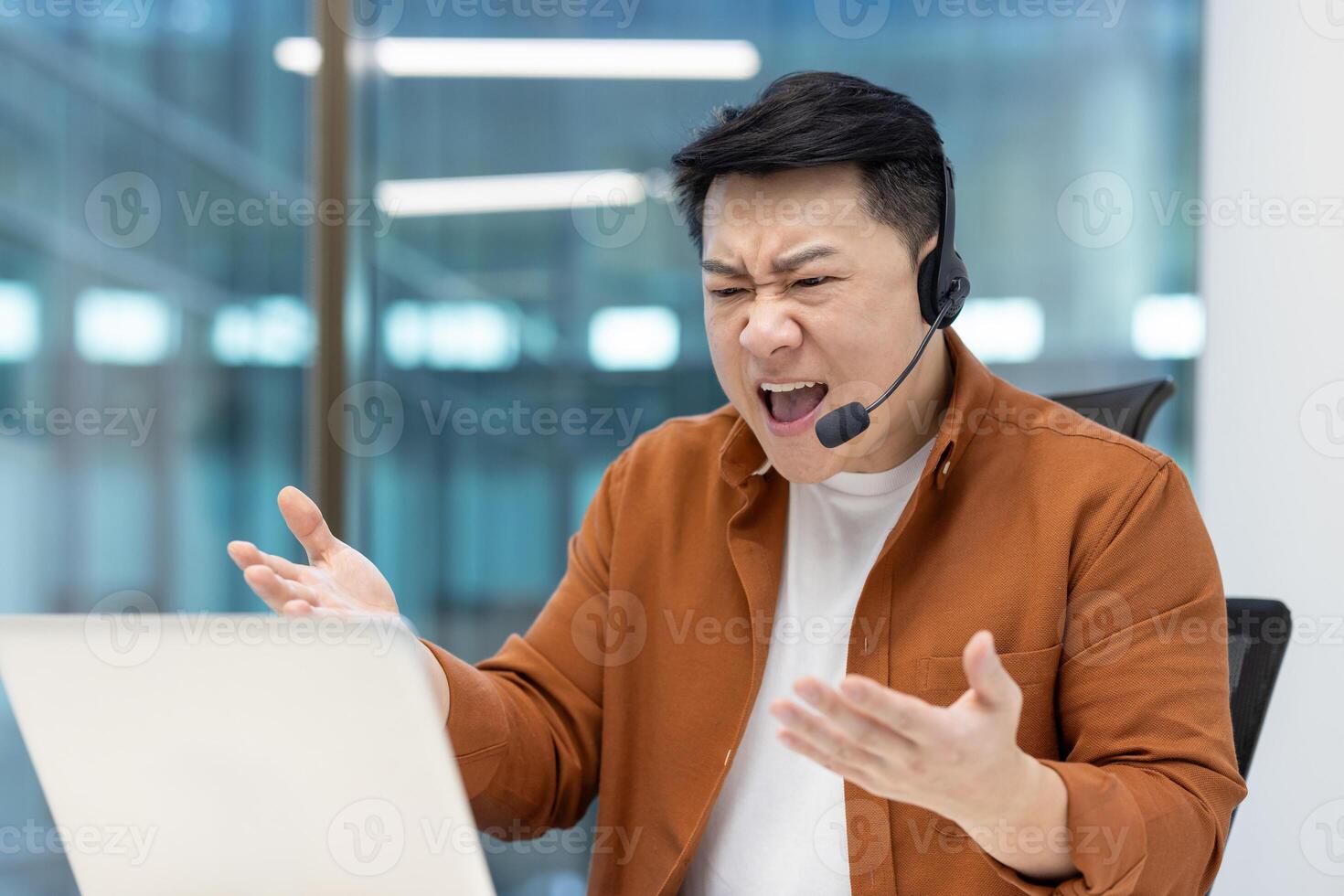 An angry Asian customer service representative is shouting at his laptop while wearing a headset in a modern office setting photo