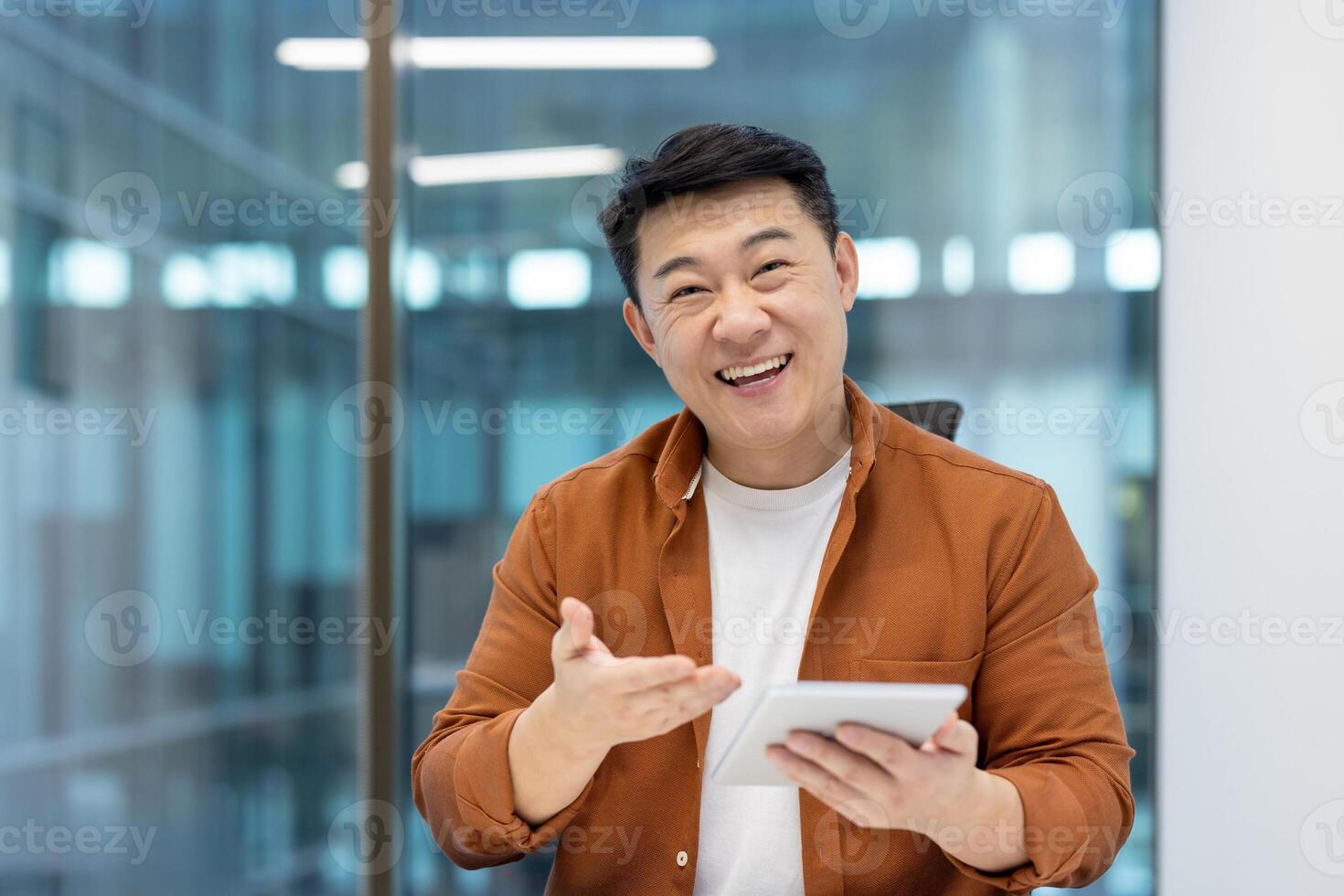 Happy asian man in a bright office setting holding a tablet and gesturing with his hand, smiling. photo