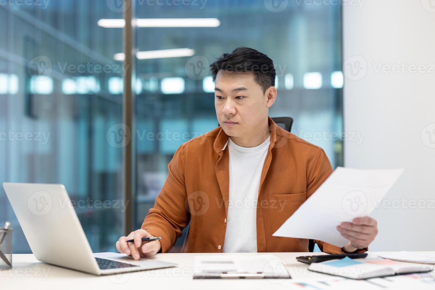 An asian businessman reviews documents while working on a laptop computer. He is seated at his desk. photo