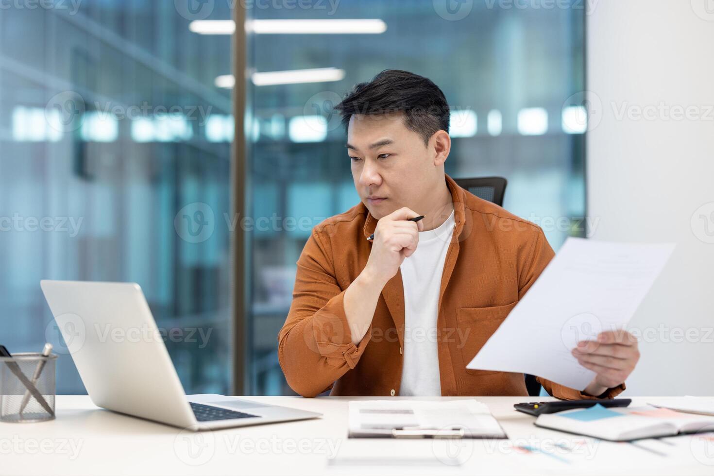 Asian businessman in office, looking thoughtfully at documents, considering data and making decisions. Analyzing business reports at his desk. photo