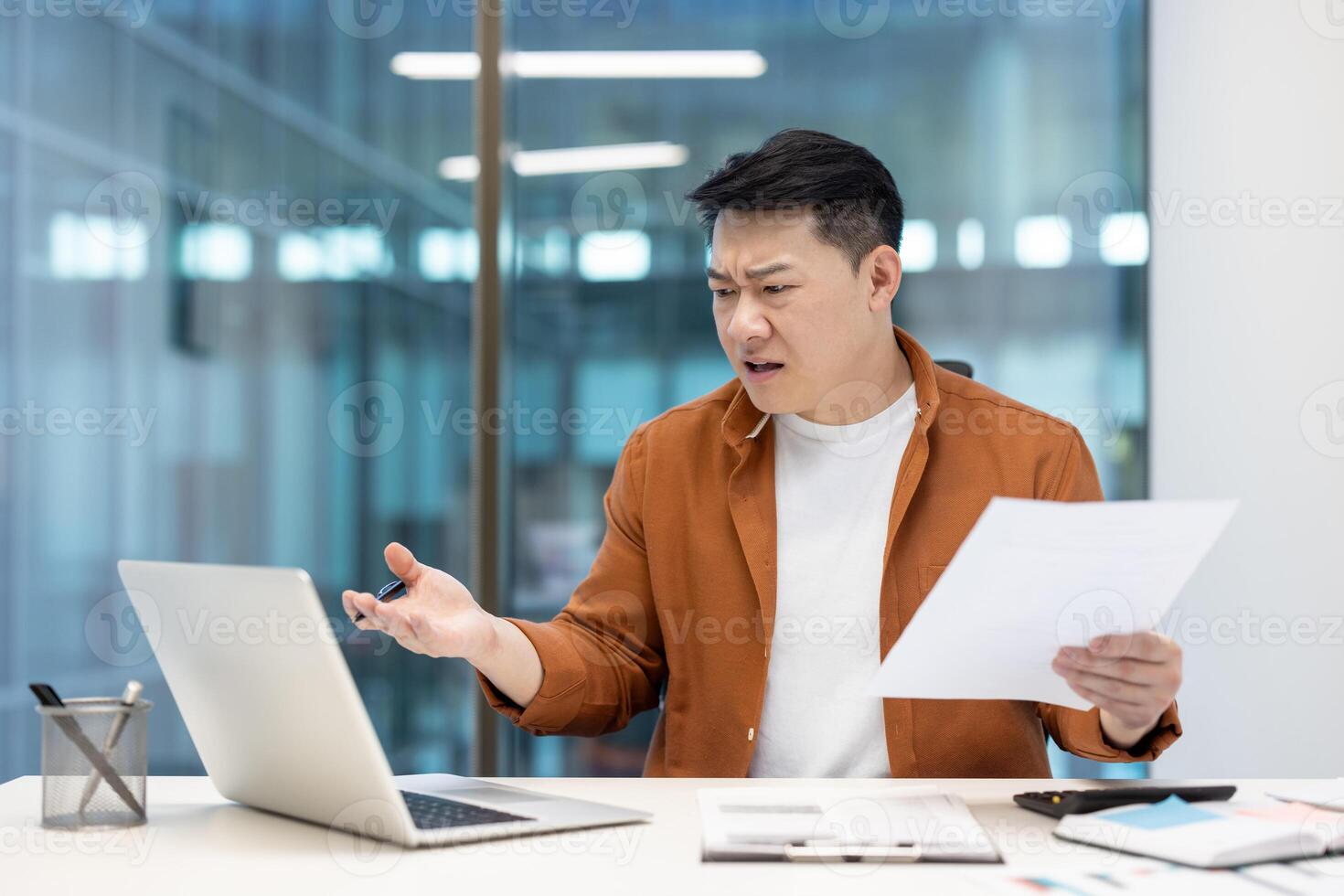Disappointed and upset financier at workplace inside office. Asian man dissatisfied with report working with documents and papers, using laptop. photo