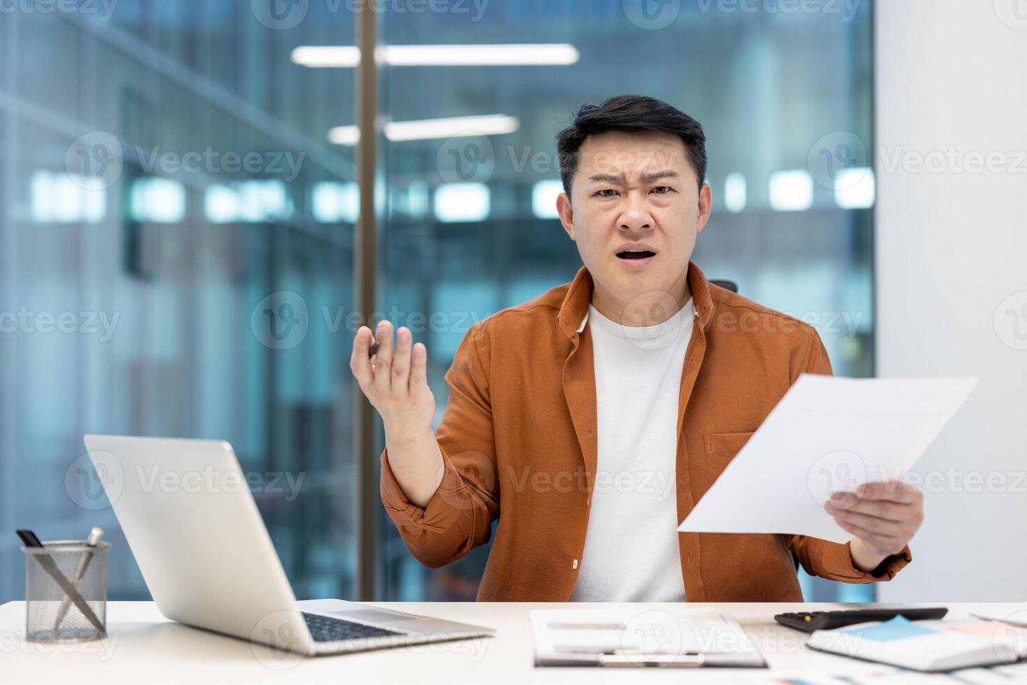 An asian businessman looks stressed holding papers, possibly reviewing documents, with a perplexed expression in a modern office. photo