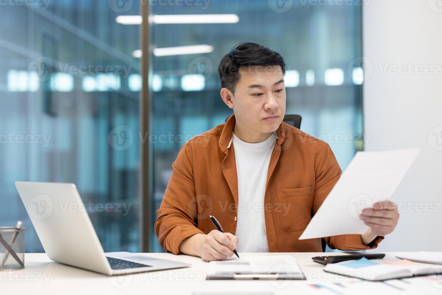 An asian businessman reviews documents at his desk in a modern office. A laptop, pen and notepad lay beside him as he works. photo