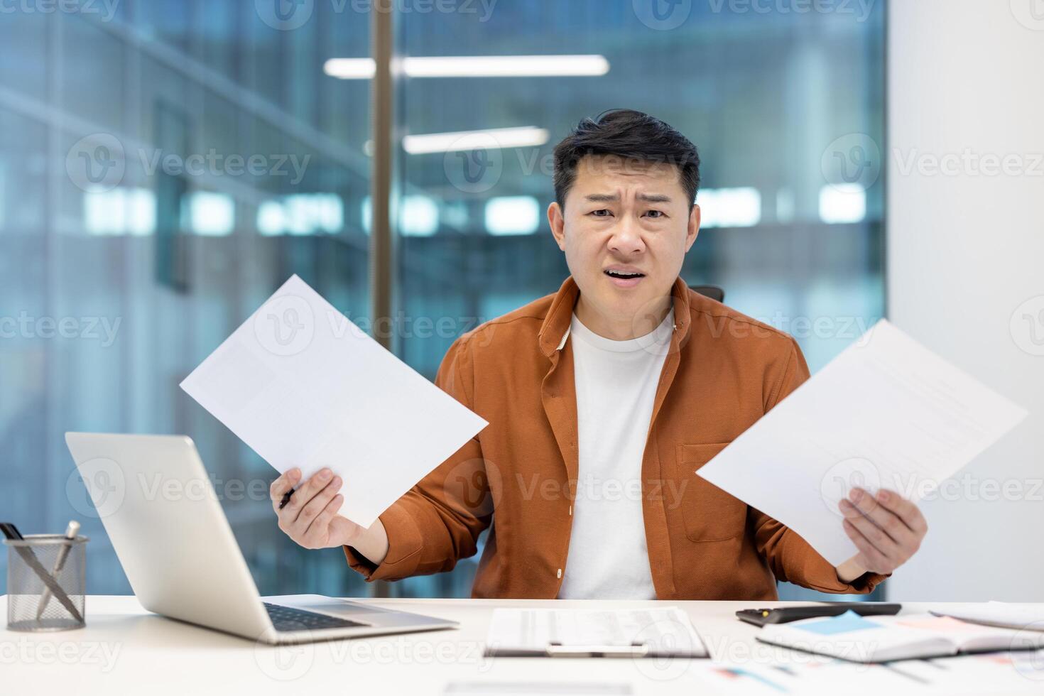 An Asian man sits at a desk in an office, holding papers with a bewildered expression, possibly struggling with work. photo