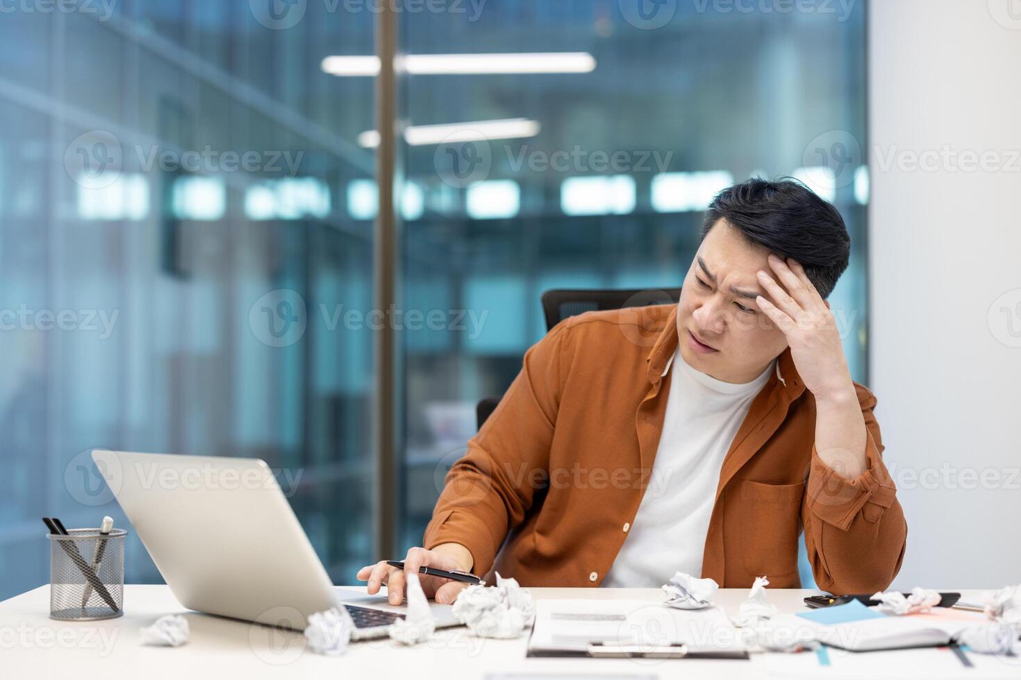An asian businessman looks stressed while working at his desk, with crumpled paper surrounding him, symbolizing writer's block. photo