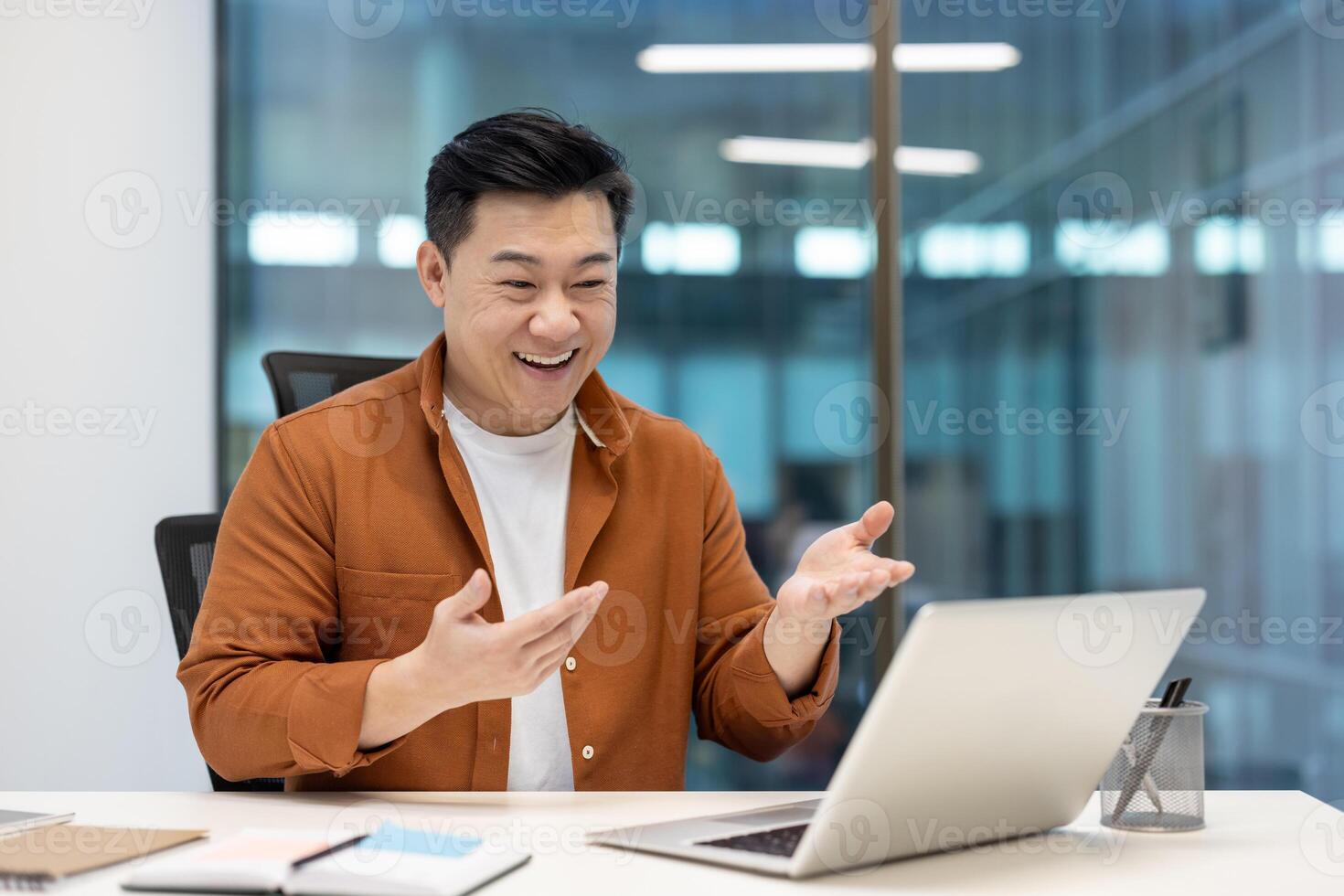 A happy asian man is sitting in an office, smiling and gesturing during a call on his laptop. Modern office setting. photo