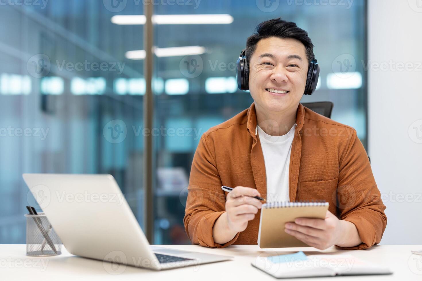 An asian businessman smiles widely while taking notes, wearing headphones, and using his laptop at work. He looks content. photo