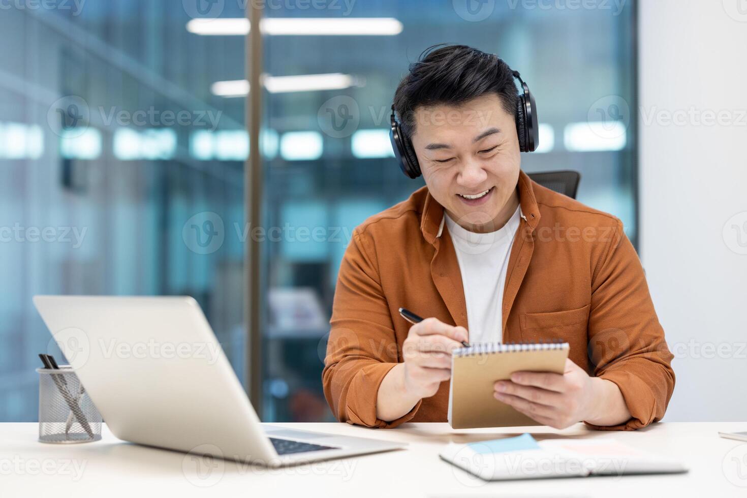 A smiling asian man wearing headphones takes notes while looking at a laptop in an office environment, working. photo
