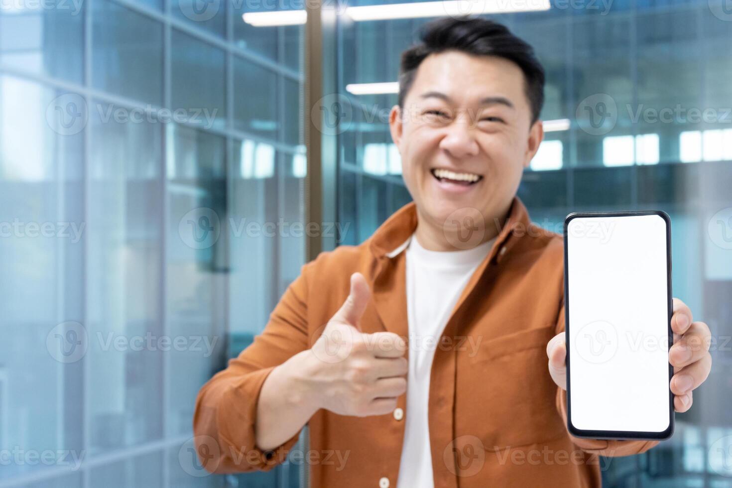 A cheerful man gives a thumbs-up while presenting a smartphone with a blank screen in a modern office environment photo