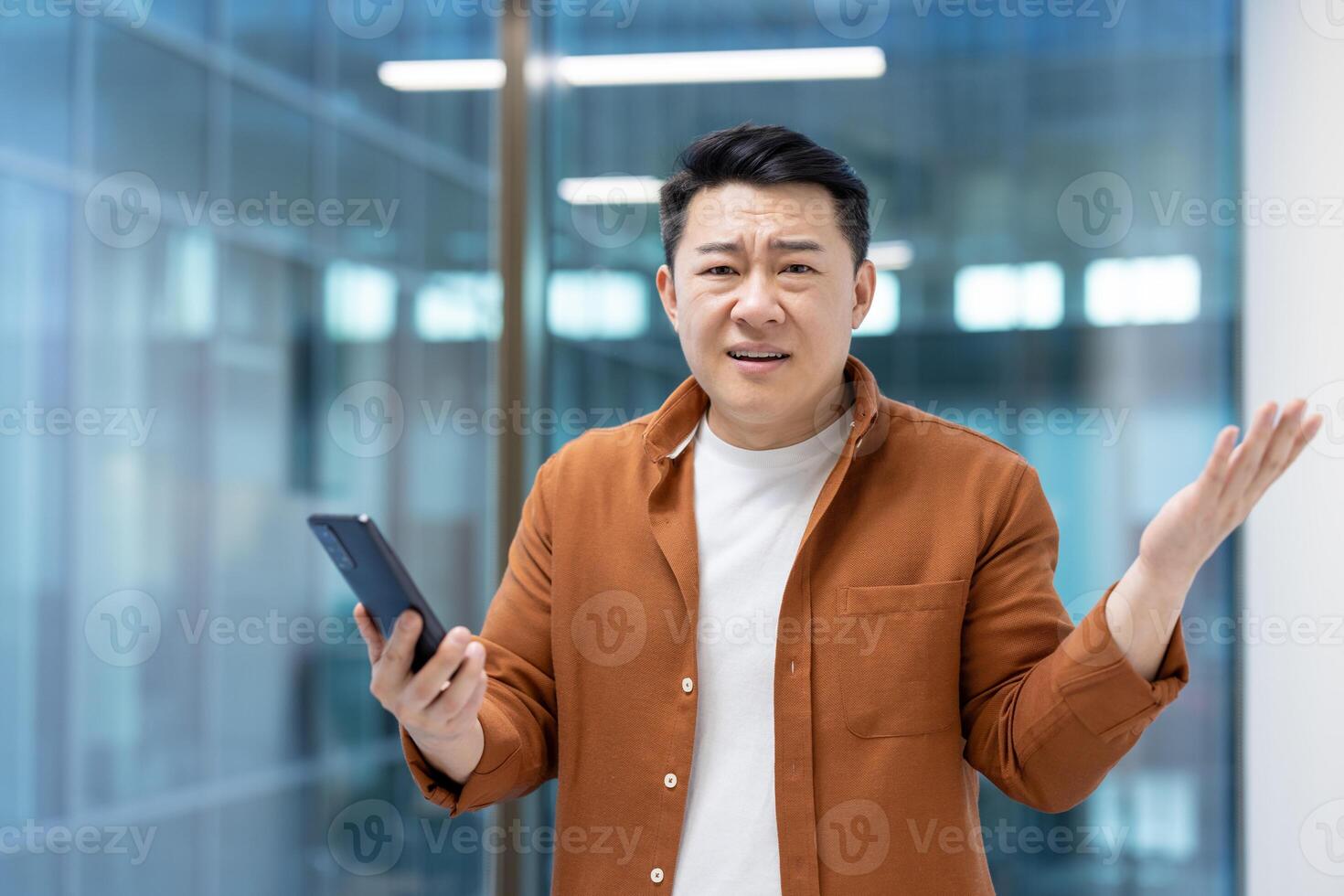 An asian man wearing an orange shirt seems to be confused by what he's reading on his mobile phone in an office setting. photo