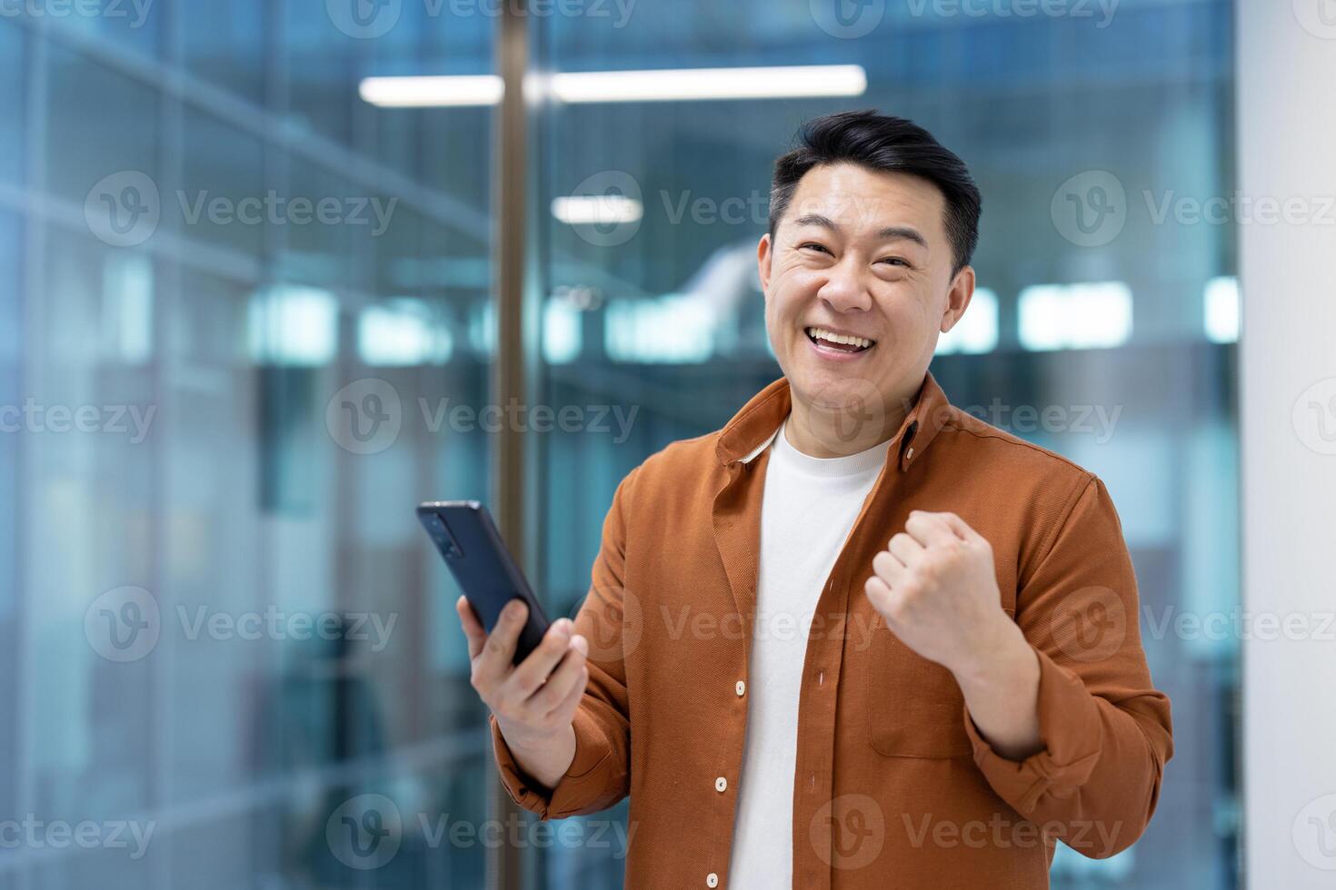 An enthusiastic asian man celebrates while holding his smartphone, showing a joyful expression. The man is inside an office setting, holding his phone with a joyful expression. photo