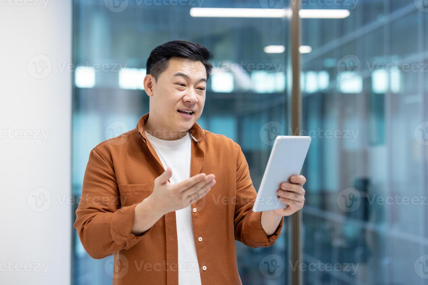 Successful Asian businessman talking to colleagues and partners using call and tablet computer. Man standing near window inside company office. photo