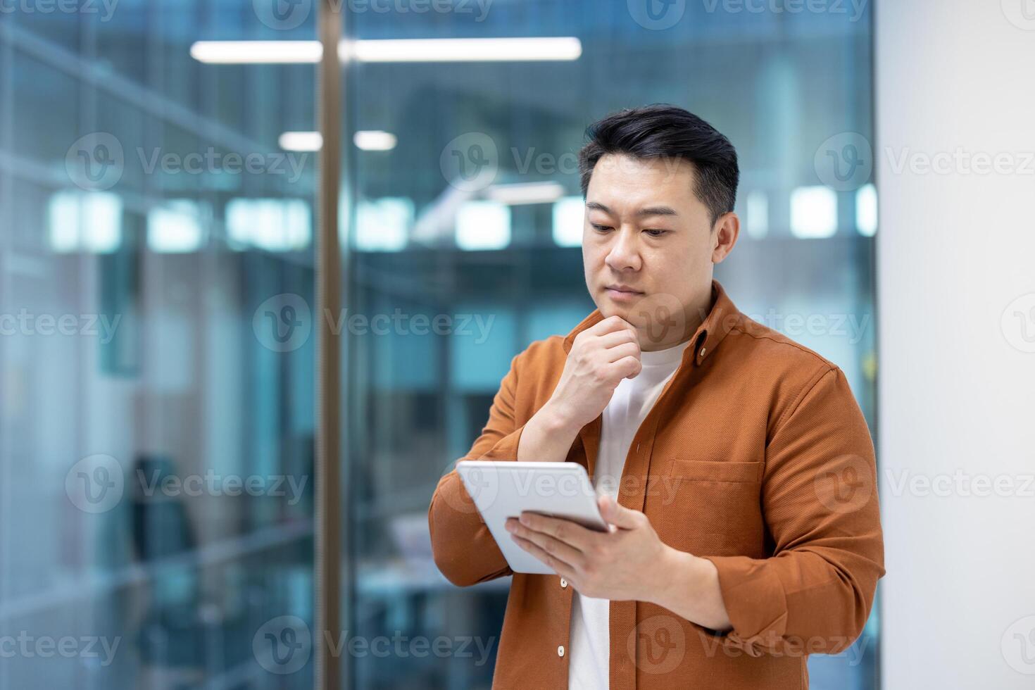 Serious thinking Asian man inside office using tablet computer. Businessman in casual clothes standing near window. photo