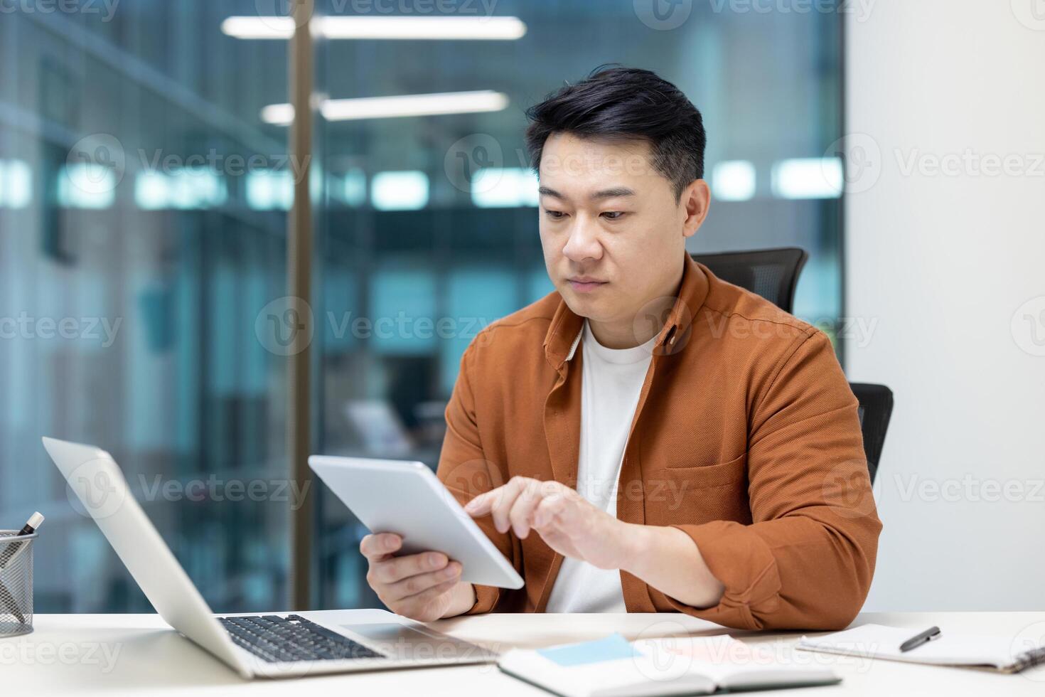 An asian businessman is seen at his desk, using a tablet. He is working on a laptop as well, focused on his tasks. photo