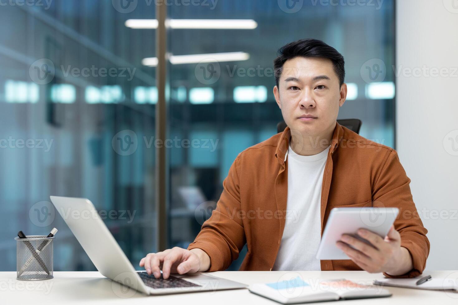 An Asian businessman is multitasking, working on his laptop while simultaneously using a tablet for a presentation in the office. photo
