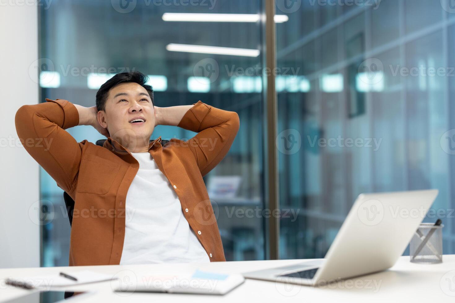 An asian professional man in an office leans back in his chair with hands behind his head, smiling and looking upwards with content. photo