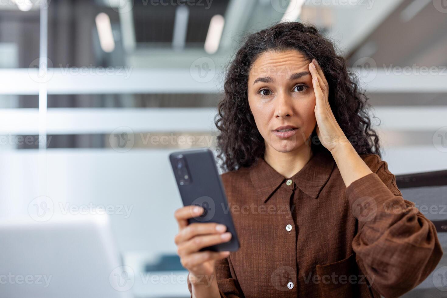 A concerned businesswoman holds her phone, looking stressed with a hand to her head in an office setting. photo