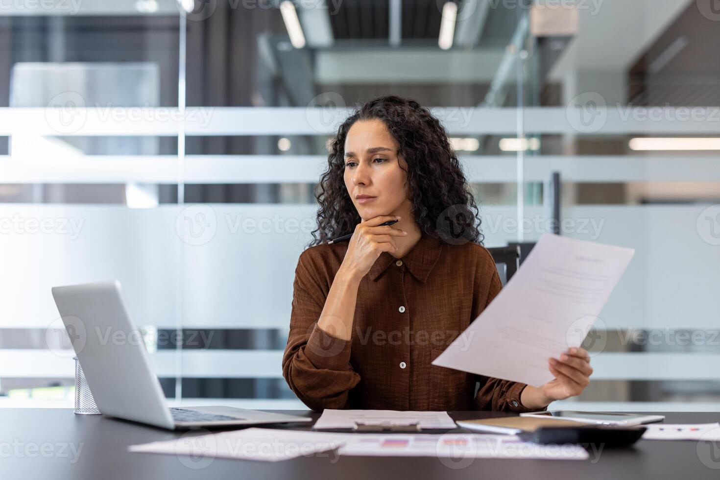 A focused businesswoman is reviewing documents and working on a laptop in a modern office setting. She appears thoughtful. photo