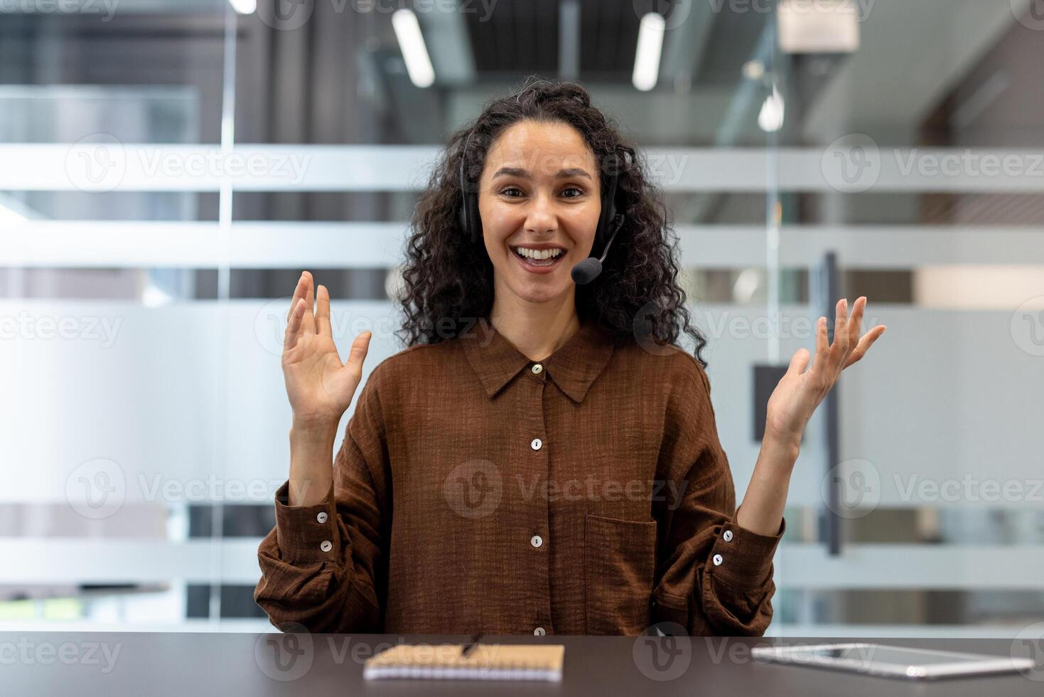 A smiling woman wearing a headset gestures while sitting at a desk in a modern office, communicating. photo