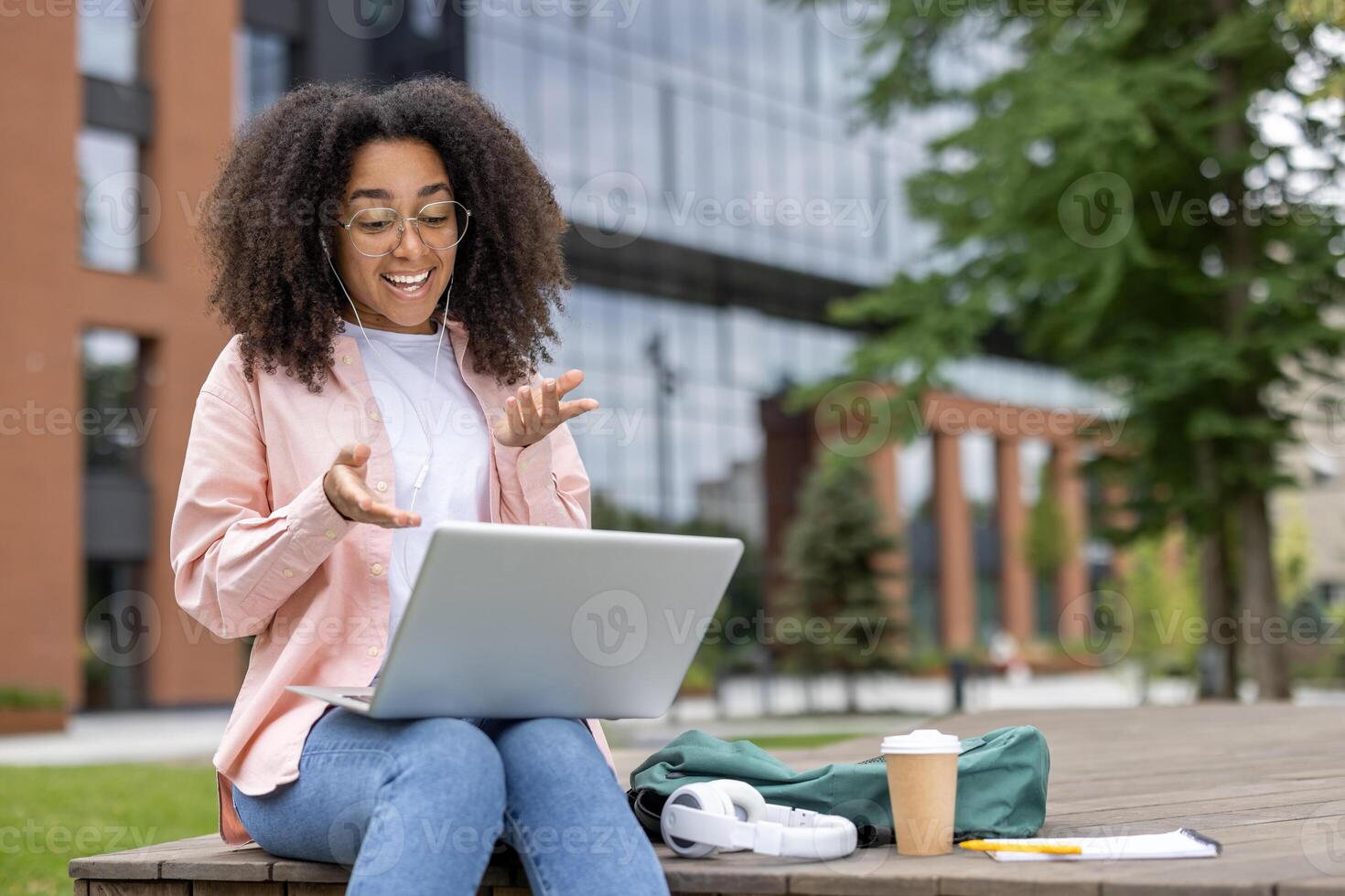 Joyful African American female student engaging in online learning and call on laptop outdoors. Relaxed and focused on communication and education with headphones and phone nearby. photo