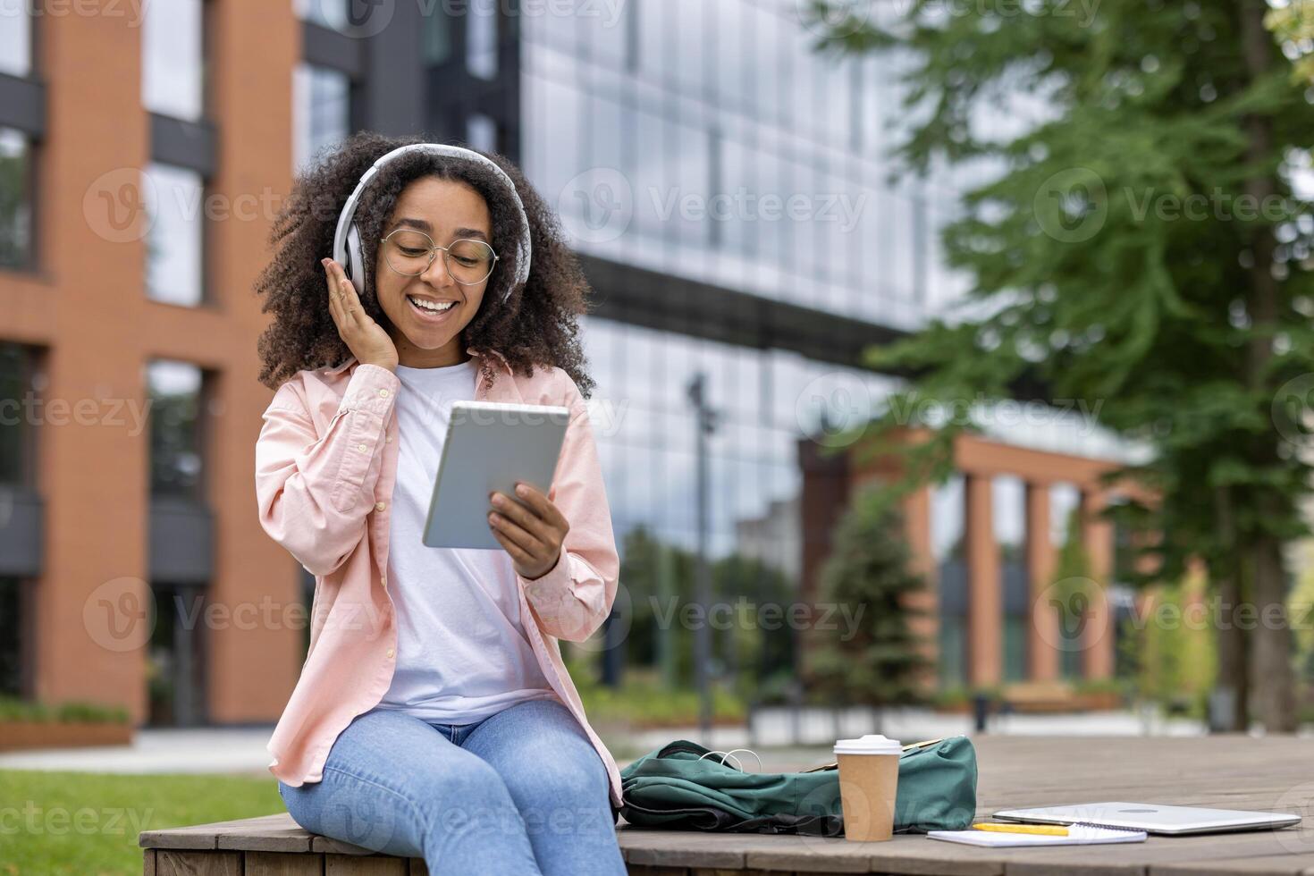 Smiling African American female student wearing headphones using tablet outside modern building. She seems relaxed and engaged, sitting on bench, enjoying music or studying. photo