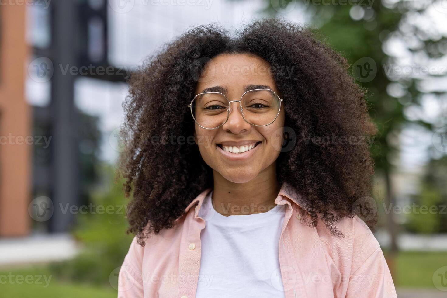 Confident African American female student with curly hair and glasses smiling warmly outdoors. Captures essence of youth and education, highlighting positive attitude and friendly demeanor photo