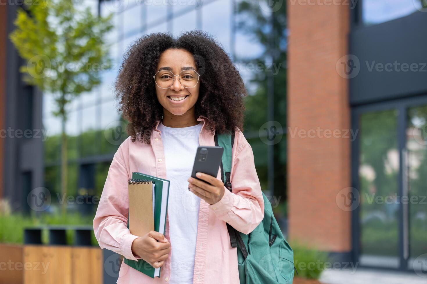 A smiling college student stands outside of a building holding a phone and notebooks. She has a backpack slung over her shoulder. photo