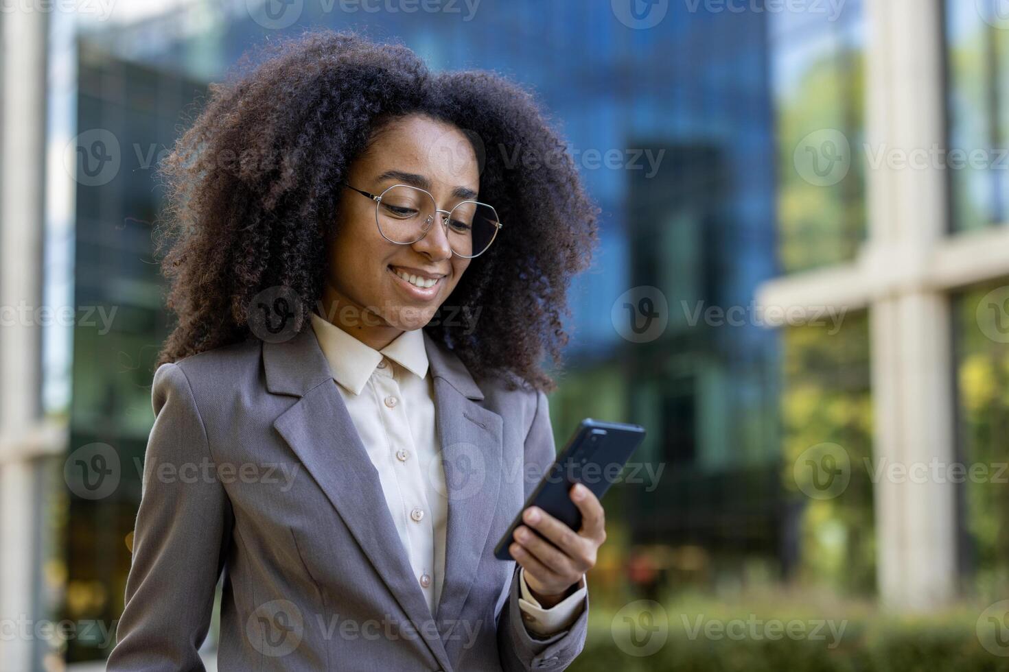 African american business woman in formal suit using smartphone outdoors. She is smiling, appearing confident and professional, standing in urban environment. Keywords. business woman, phone. photo