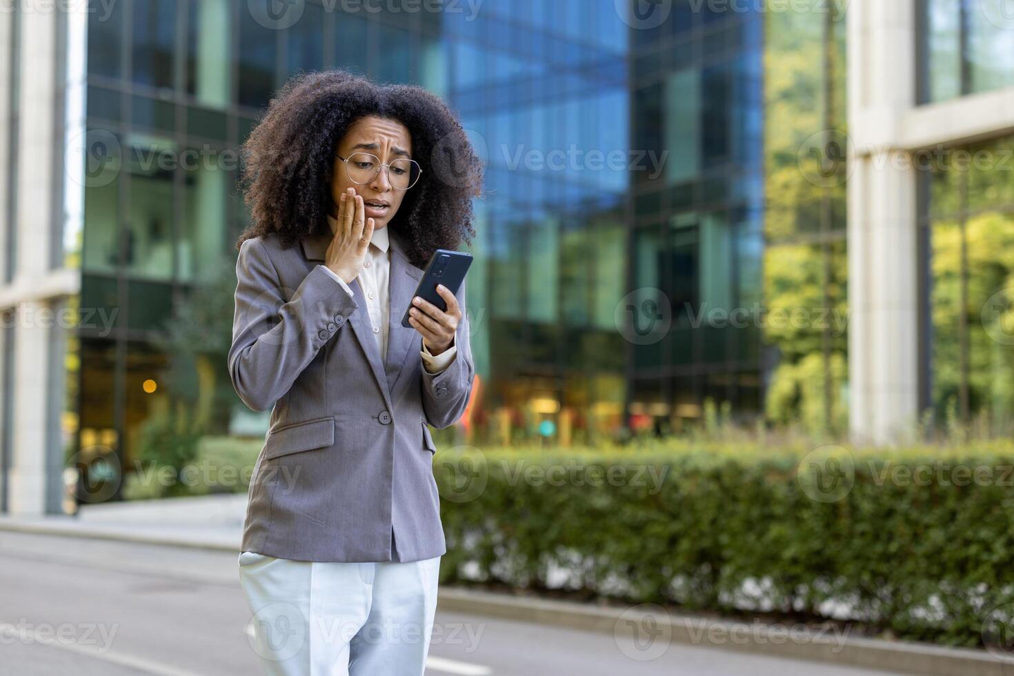 Hispanic businesswoman in formal suit standing outside office building, holding phone with surprised expression. Captures professional setting, unexpected news, and urban environment. photo