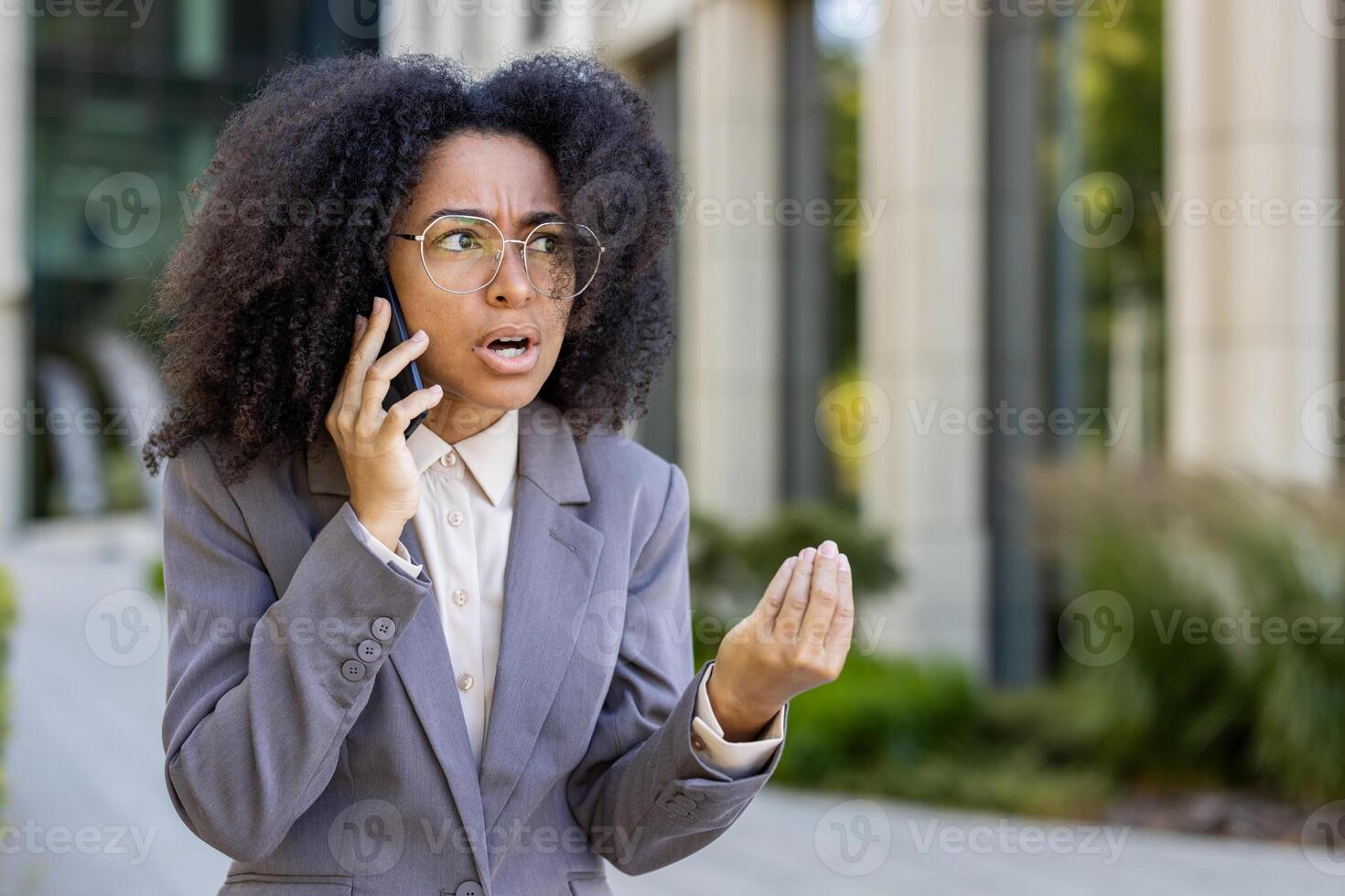 Professional african american business woman in gray blazer engaged in tense phone conversation. Her expressive hand gestures and worried expression convey frustration, urgency, and business stress. photo