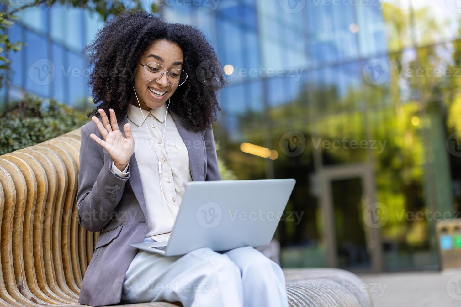 Smiling African American business woman using laptop for call in park. Wearing glasses and suit, she waves at screen, expressing positivity. Technology, remote work, and modern communication photo