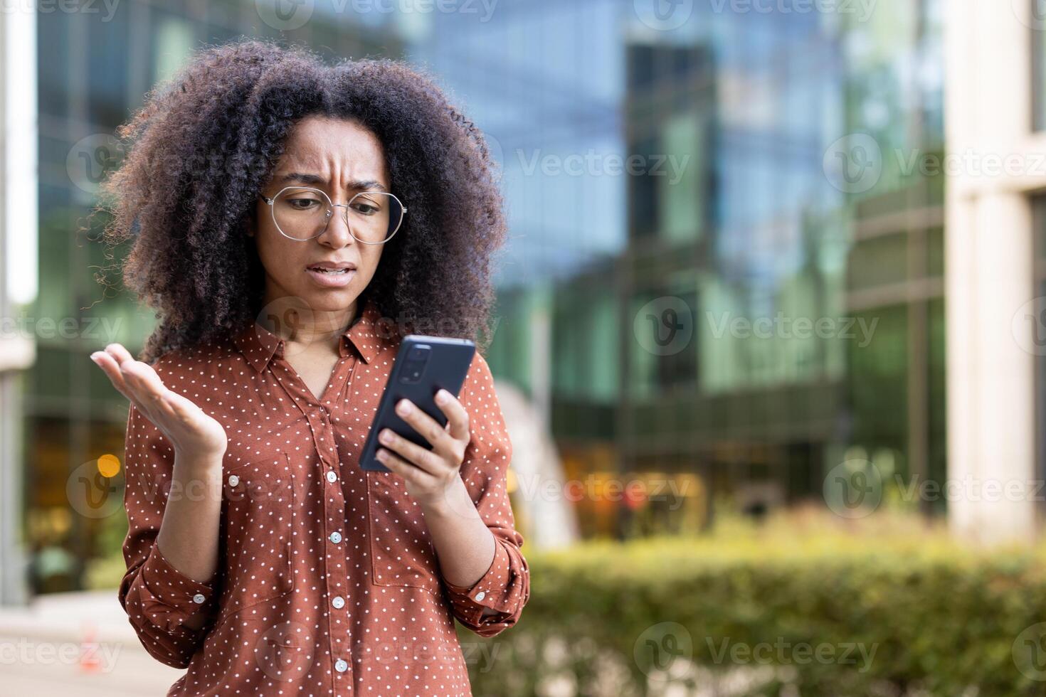 A frustrated woman in glasses looks at her smartphone, seemingly upset with the content she is viewing. The background is a blurred cityscape. photo