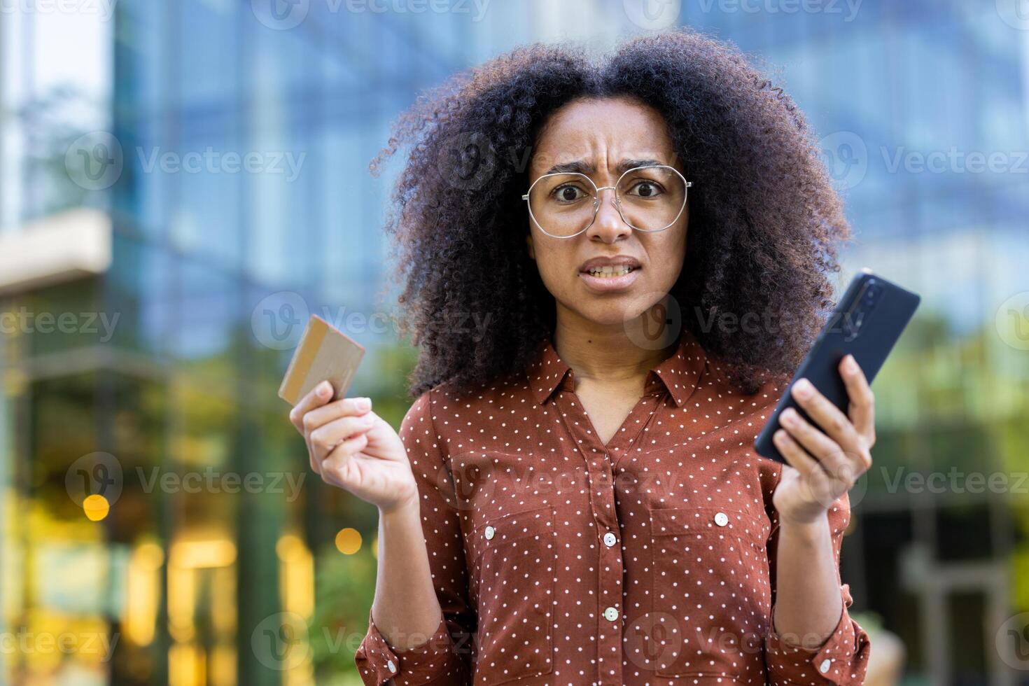 A frustrated woman with glasses holds a credit card and smartphone, possibly experiencing an issue. The setting includes an office building. photo