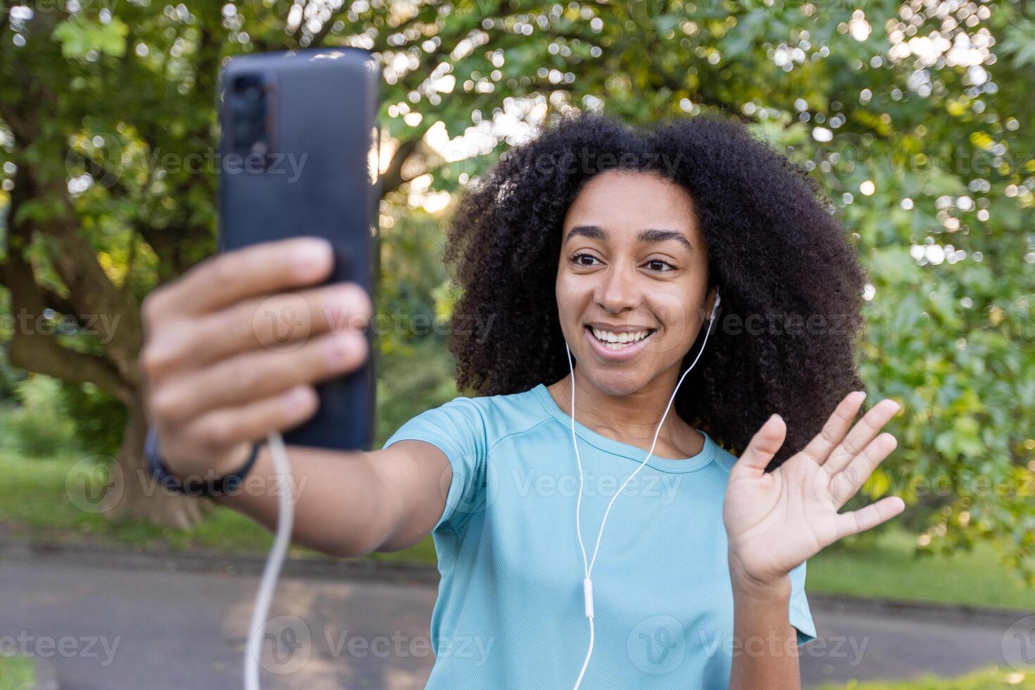African American woman enjoying outdoor call using smartphone. She is smiling and waving while wearing earphones. Captures feelings of connection, happiness, and technology a natural setting. photo